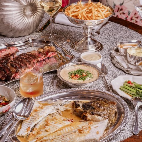 A table full of food at Lilibet's Mayfair, including fish, steak, and vegetables on silver plates and platters