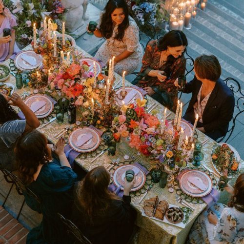 An aerial view of people eating at a rectangular table decorated with pink flowers, plates, and candles